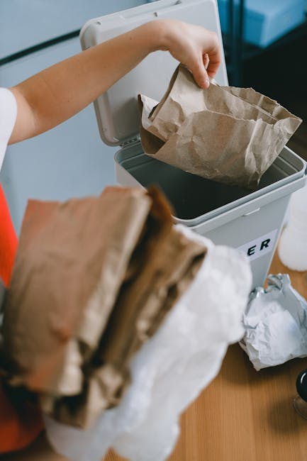 A person's arm is reaching into an open grey recycling bin with a black lid, lifting a crumpled brown paper bag. In the foreground, another crumpled brown paper bag is being held, partially obscuring 